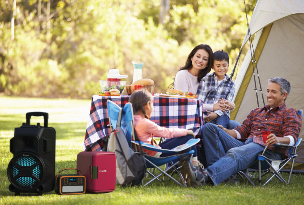 A family on a camping trip together, as a tzumi MegaBass Speaker, ionchill Mini Fridge, and tzumi Vault Power Station sit next to them.